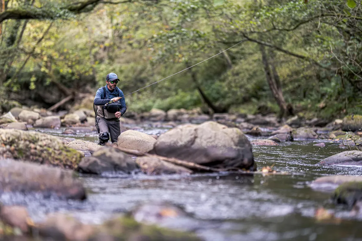 Angler standing in a clear river flowing over boulders through green forest