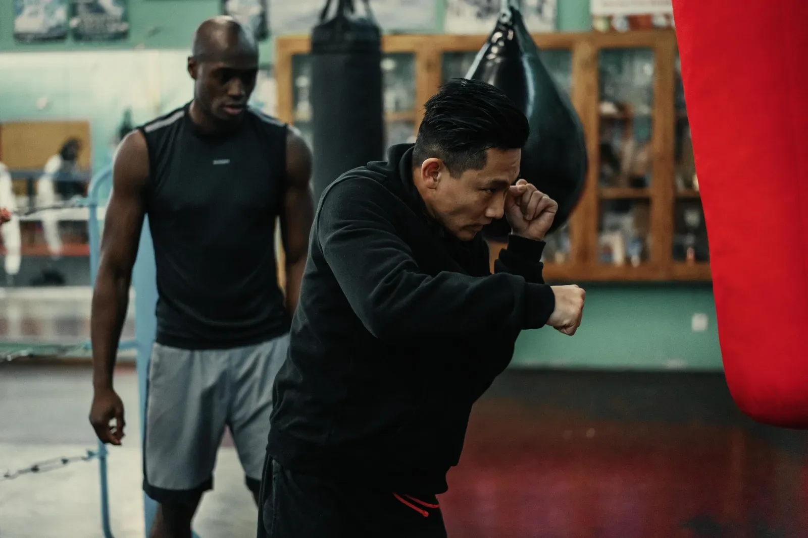 Boxing coach watching a fighter hold his guard during a drill beside a red heavy bag