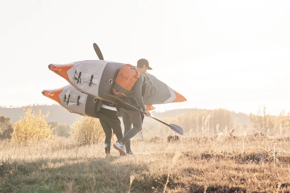 Two people carrying inflatable kayaks across golden grass at sunrise