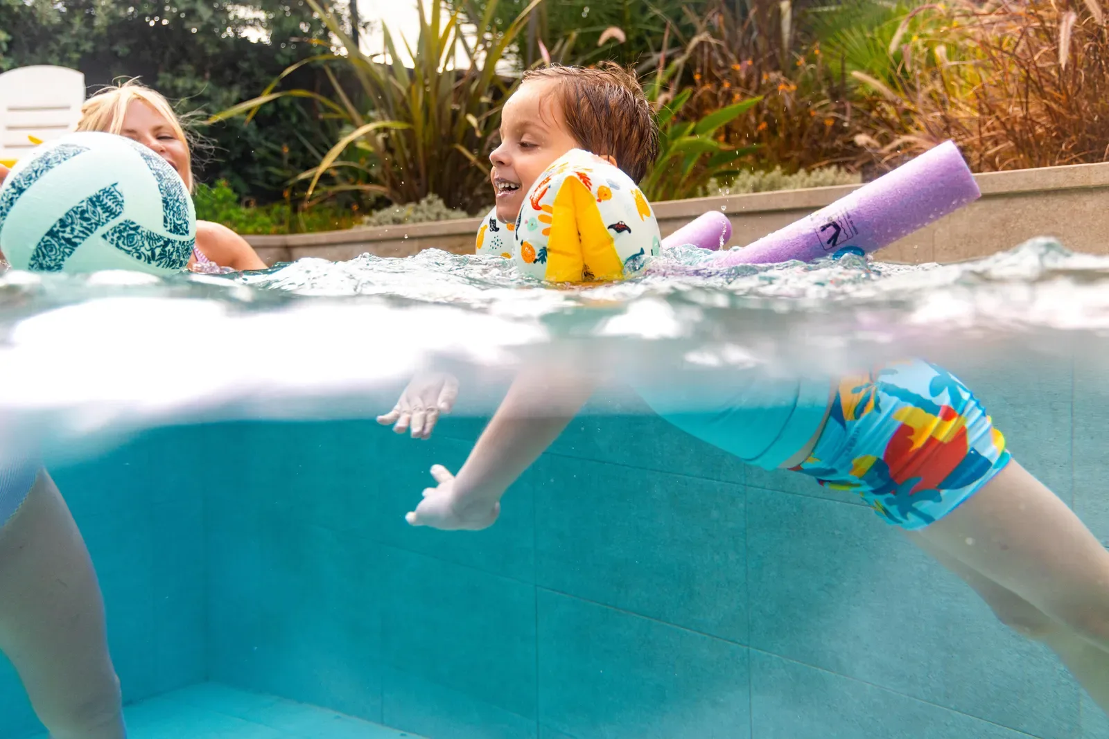 Young child in armbands kicking in a pool with a parent, a beach ball and pool noodles nearby