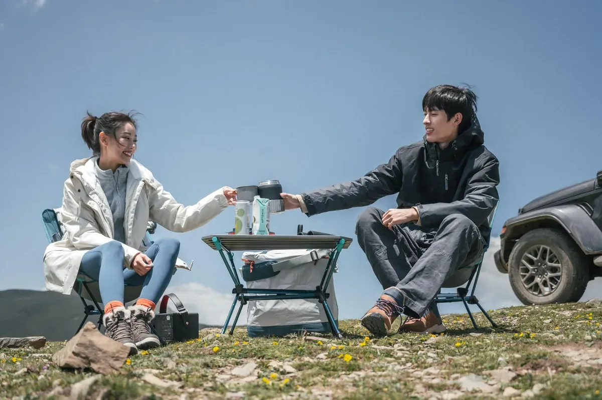 Couple in warm jackets sharing a hot drink at a camp table beside their 4WD on a high meadow