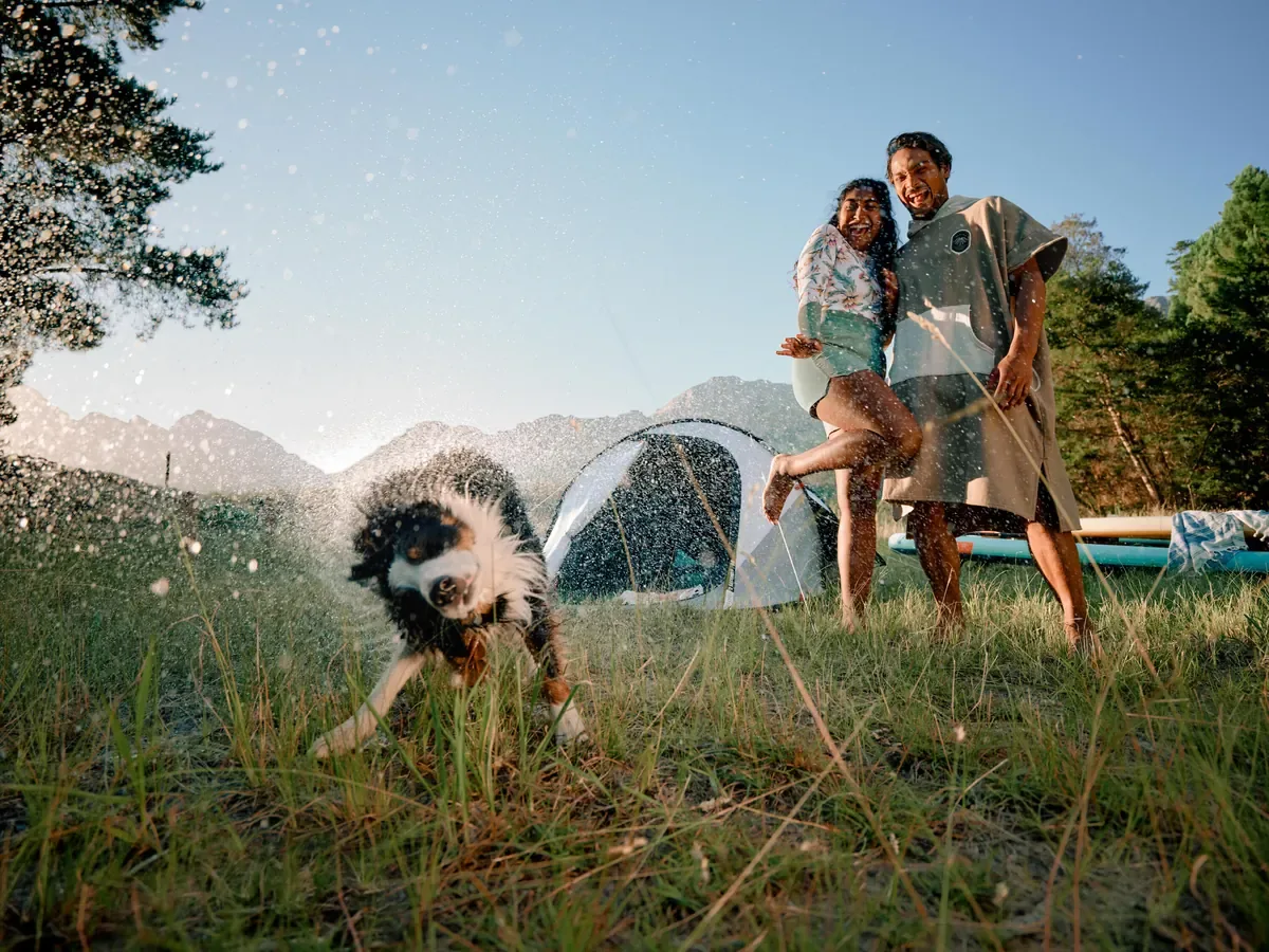Wet dog shaking off water on a grassy campsite while a couple laughs beside their tent