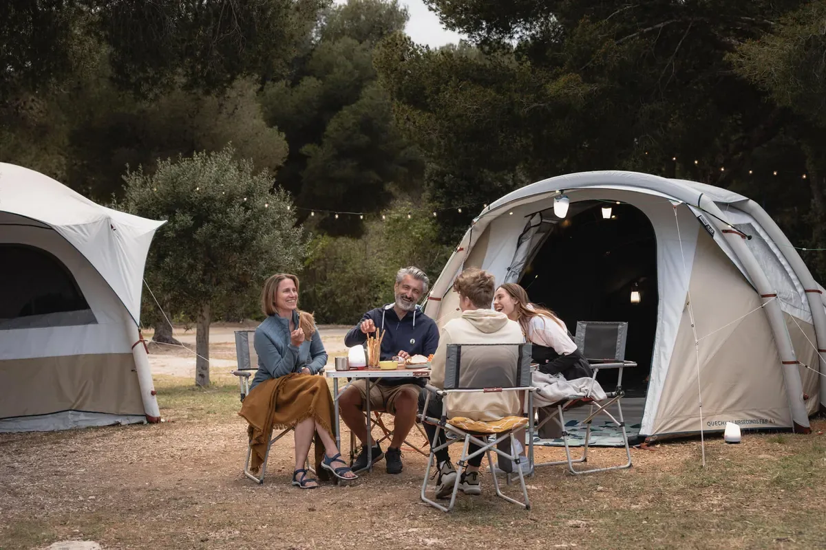Family sharing an evening meal at a camp table between tents under string lights