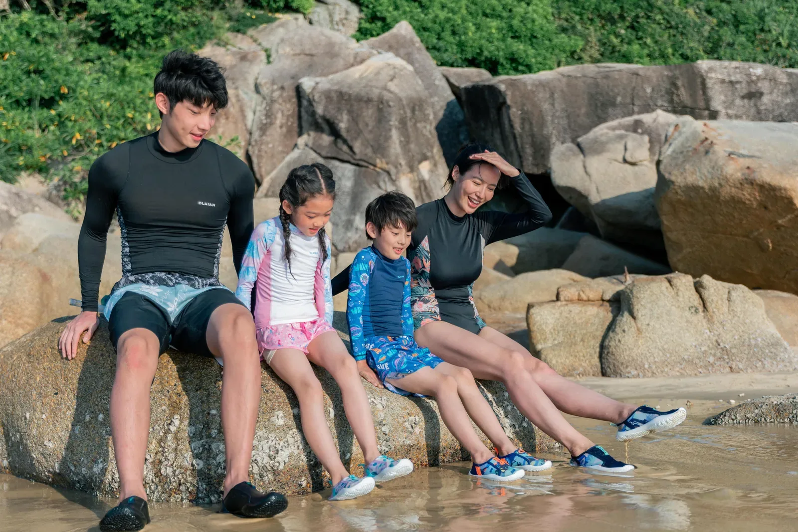 Family in rash vests and water shoes sitting together on rocks beside shallow water
