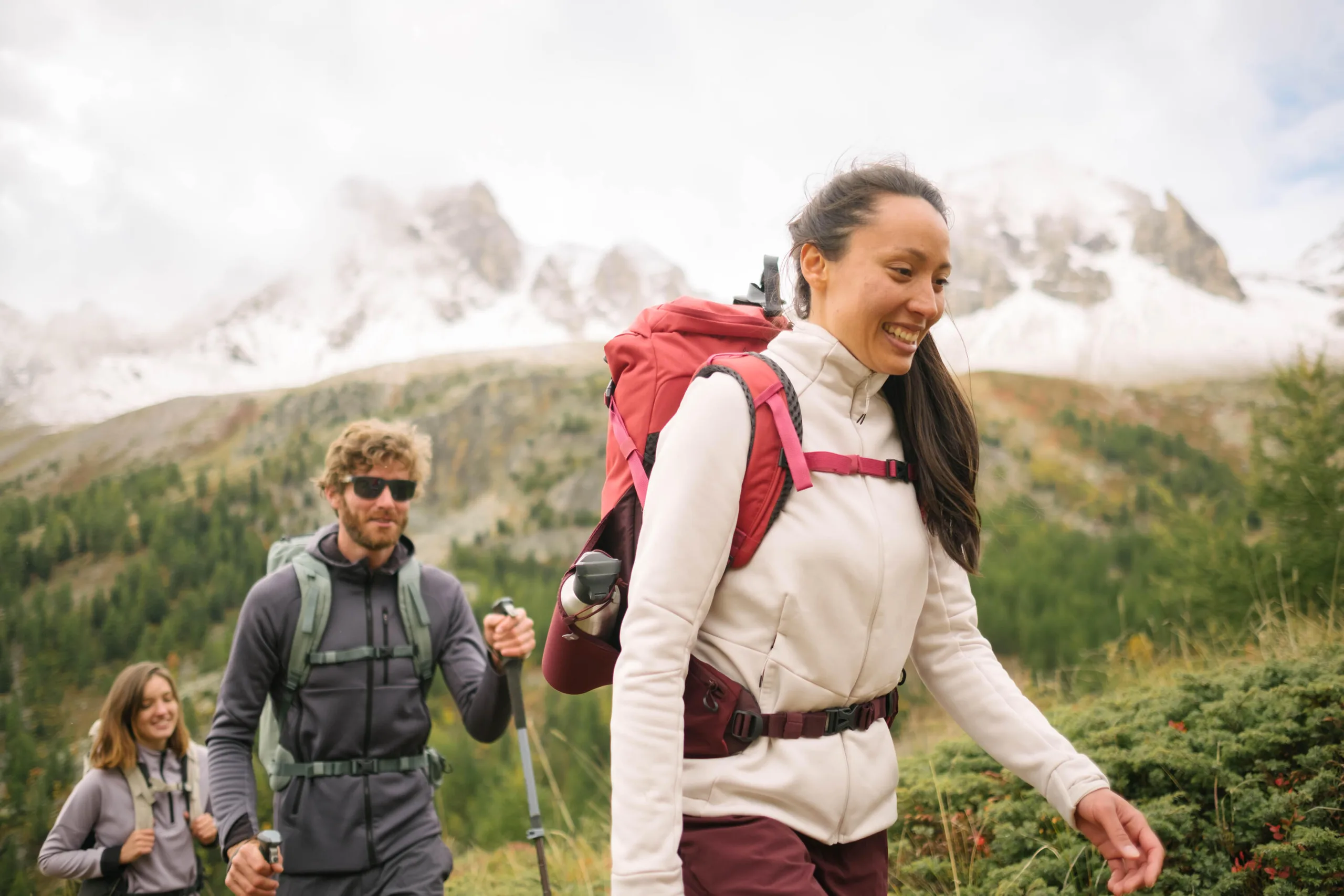 Three friends with backpacks and poles hiking through a green alpine meadow