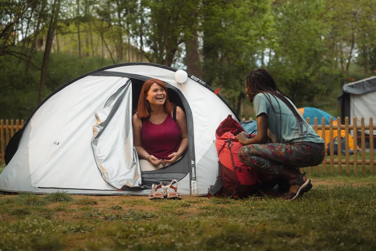 Friend smiling from the doorway of a pop up tent while another unpacks a backpack at a grassy campsite
