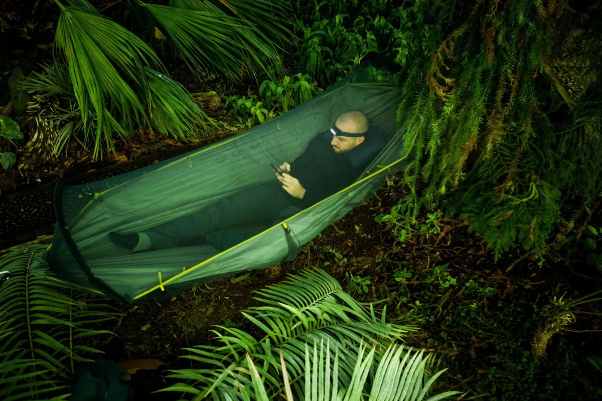 Camper resting in a hammock tent strung among lush ferns in green forest