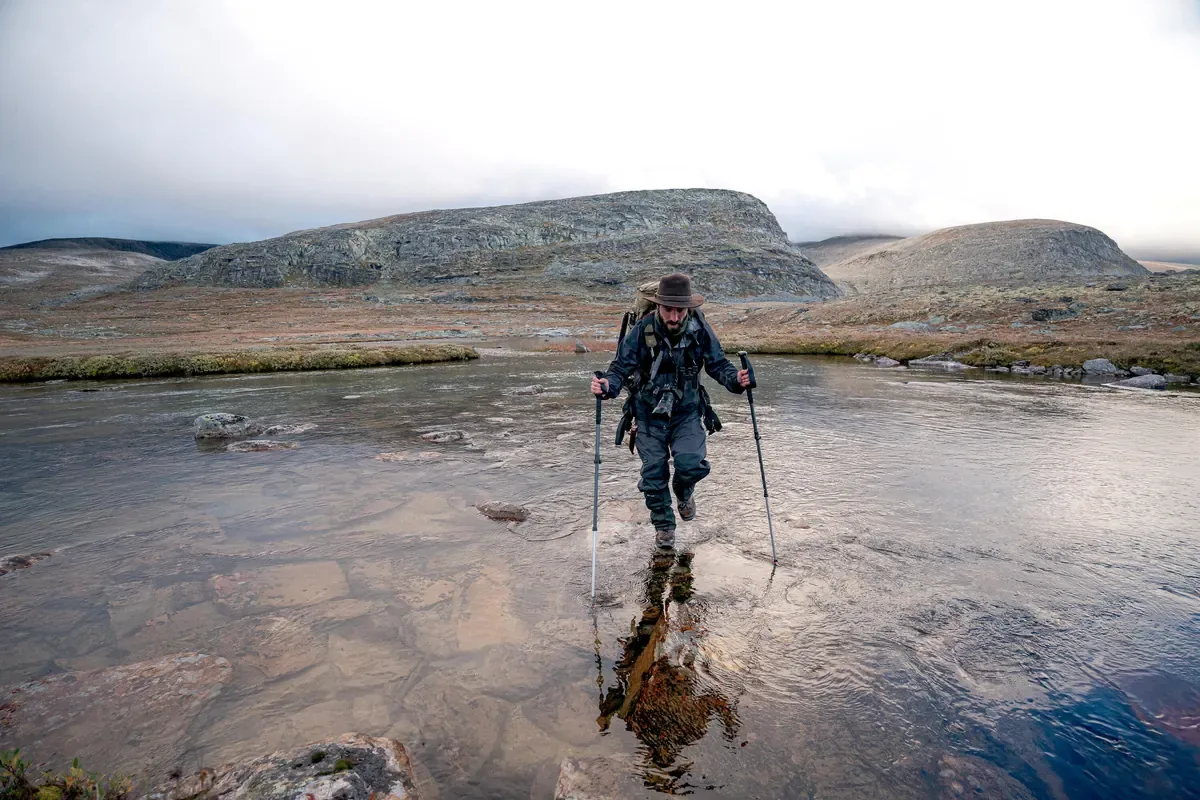 Hiker with trekking poles wading across a shallow river on an open high plain