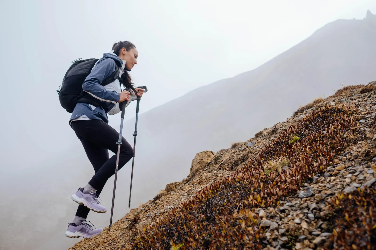 Hiker with trekking poles climbing a steep slope with misty mountains behind