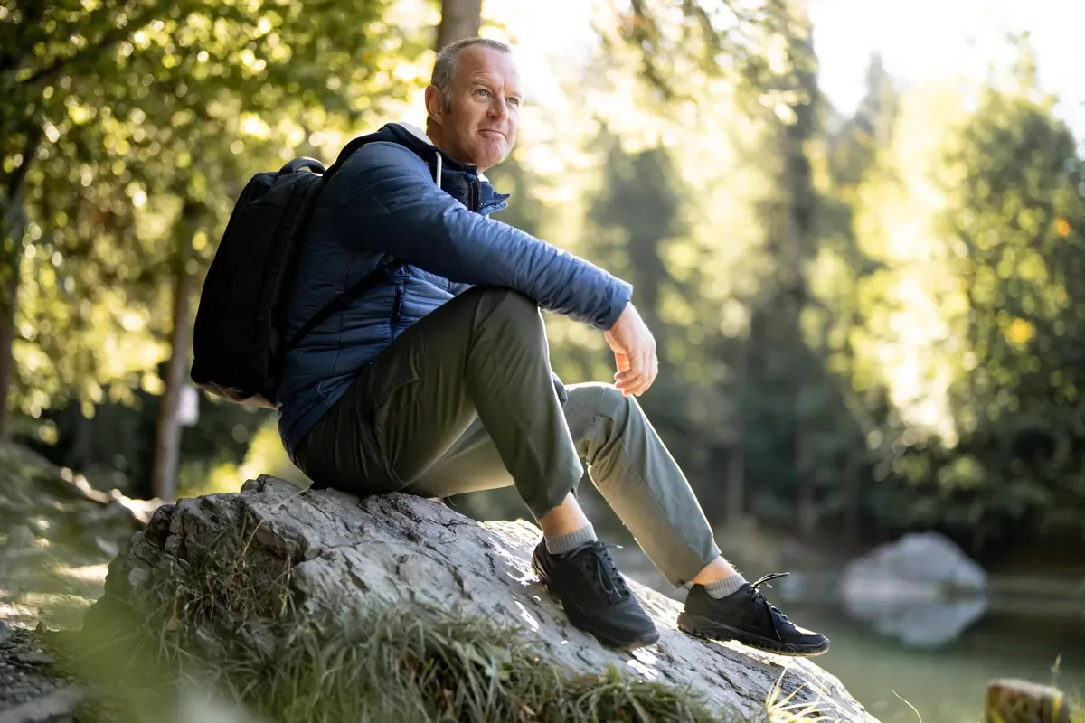 Hiker with a daypack resting on a boulder beside forest and water in golden light