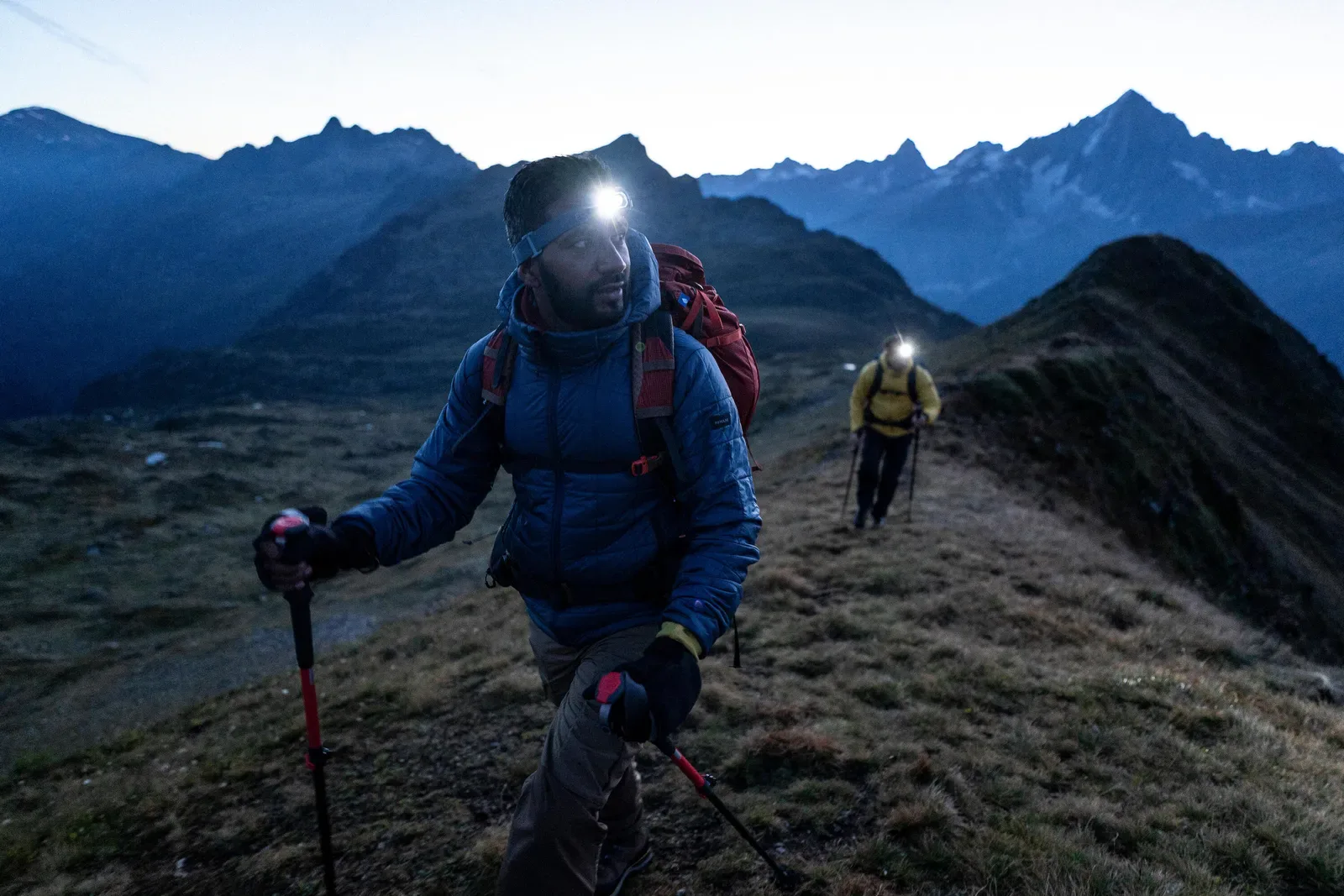 Two hikers with head torches and trekking poles on a grassy ridge at dusk