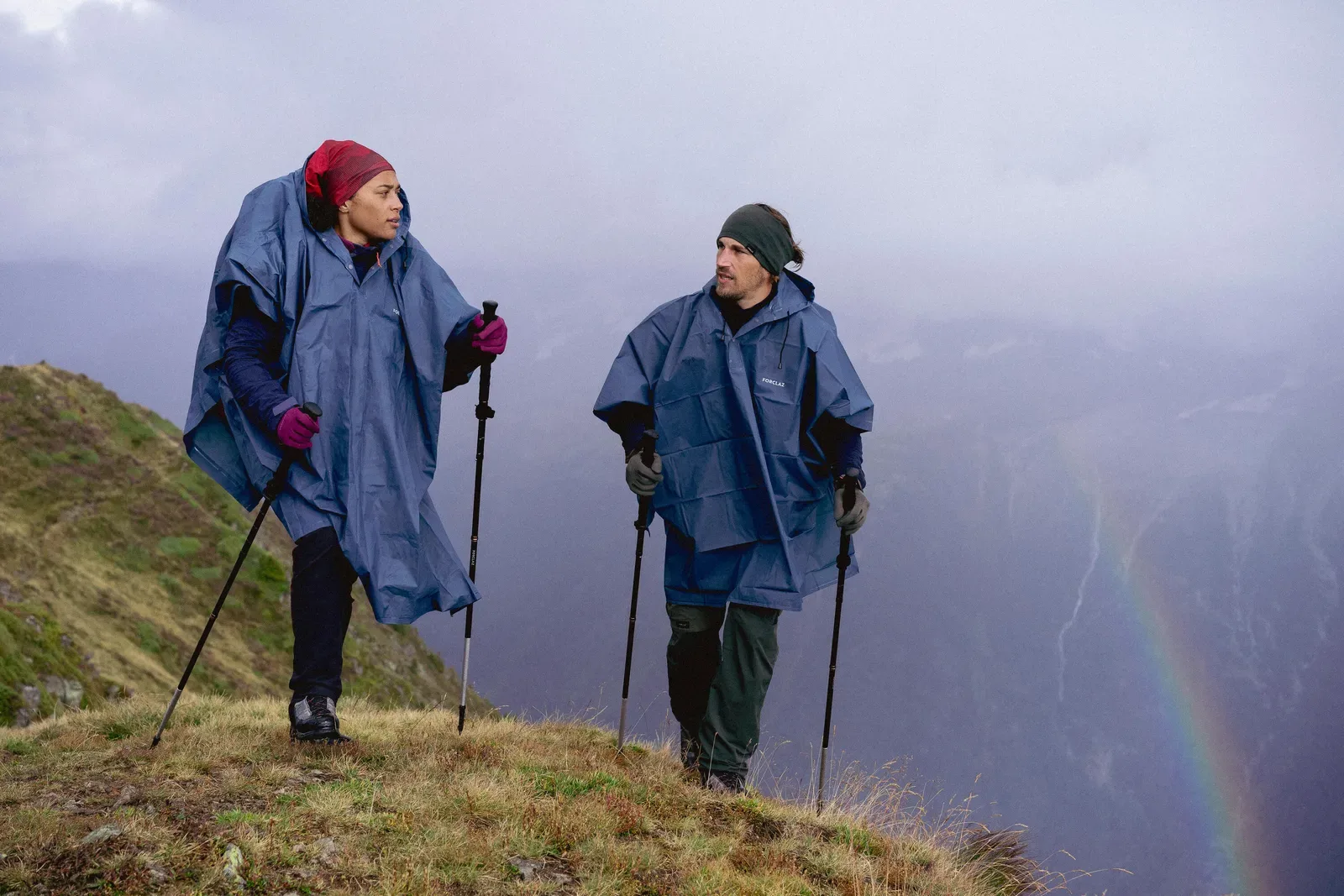 Two hikers in rain ponchos with trekking poles on a grassy ridge, a rainbow in the valley below