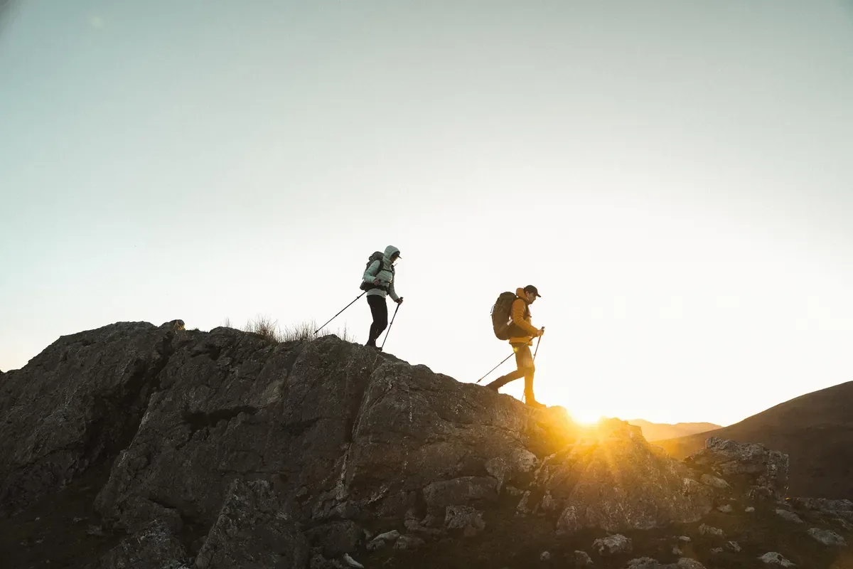 Two hikers with packs and trekking poles silhouetted on a rocky ridge at sunset