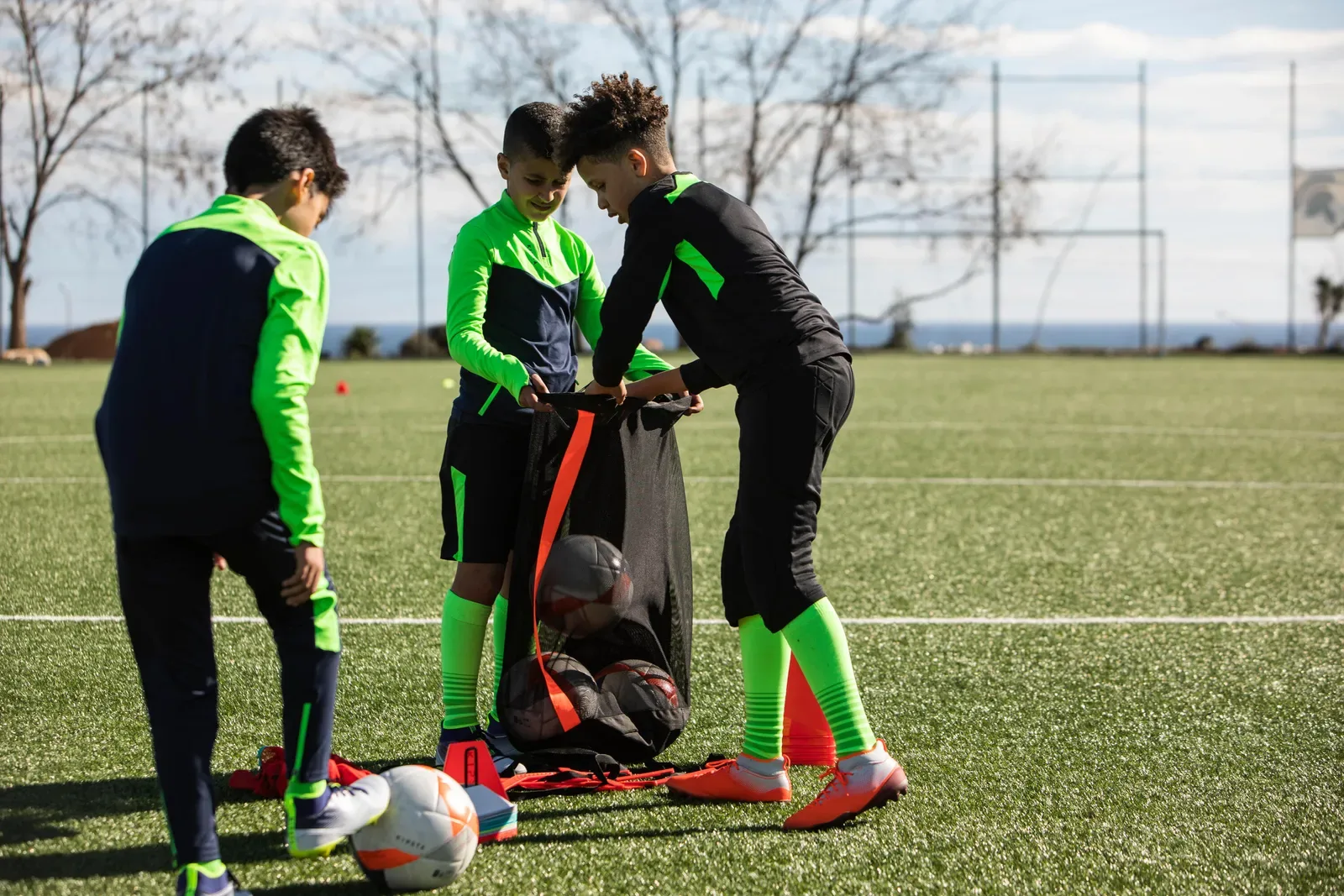 Three junior soccer players packing balls into a mesh ball bag on the pitch