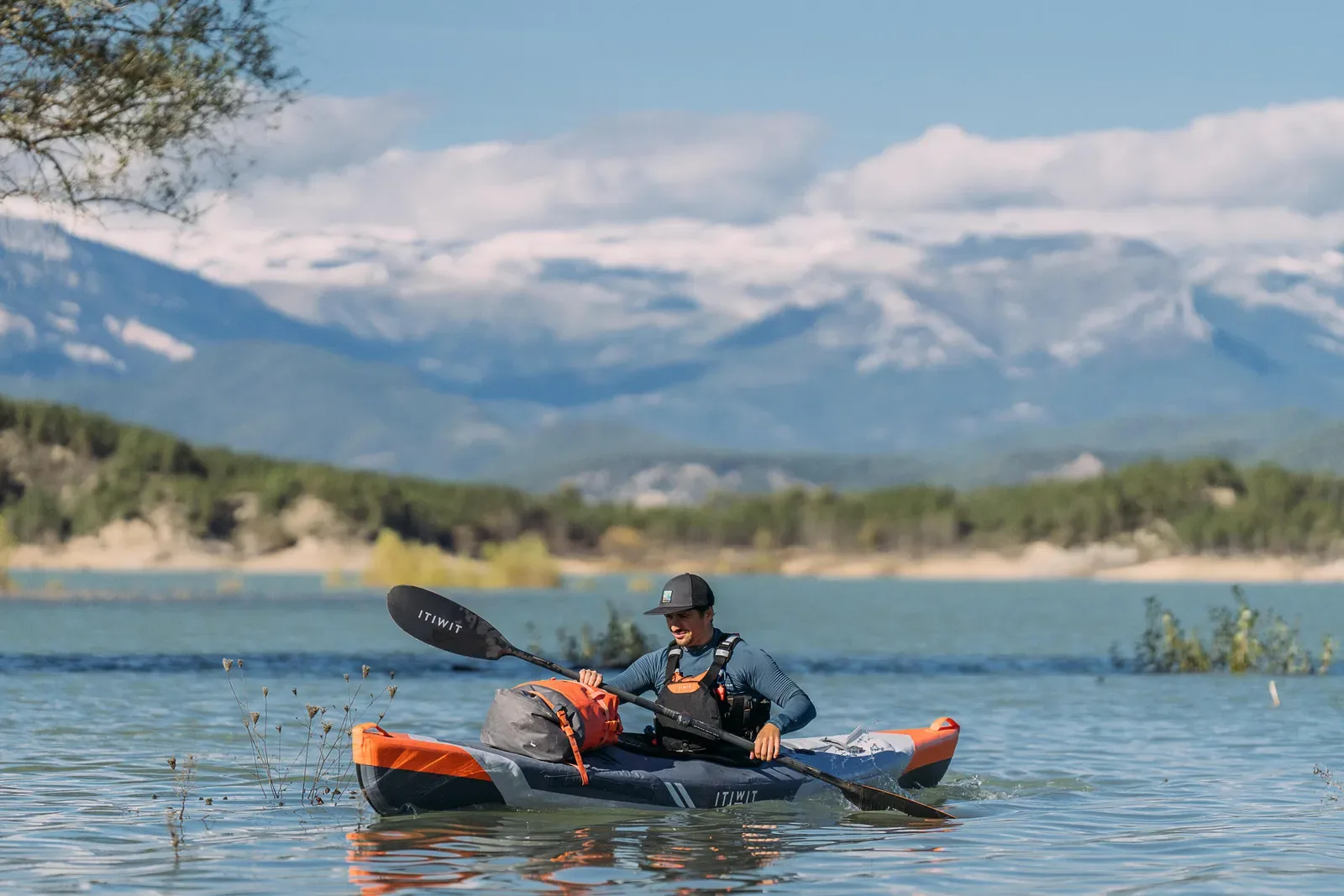 Man paddling an inflatable kayak on a calm lake with snowy mountains behind
