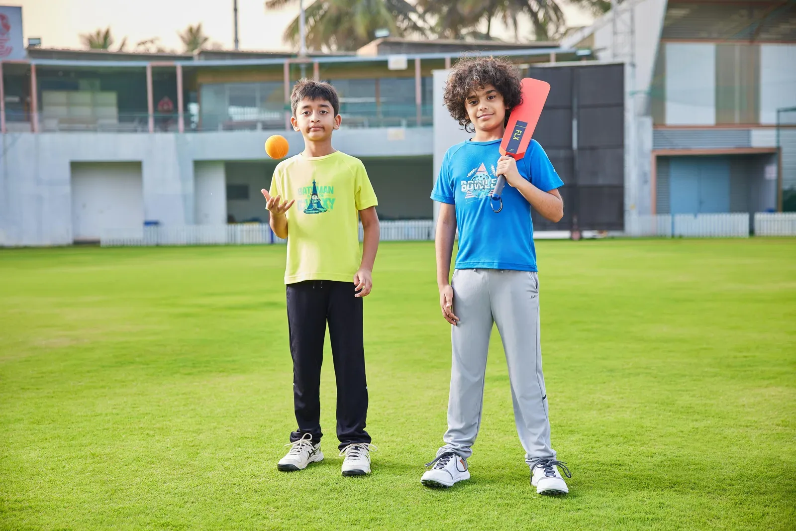 Two kids on a cricket oval, one tossing an orange ball, one holding a bat