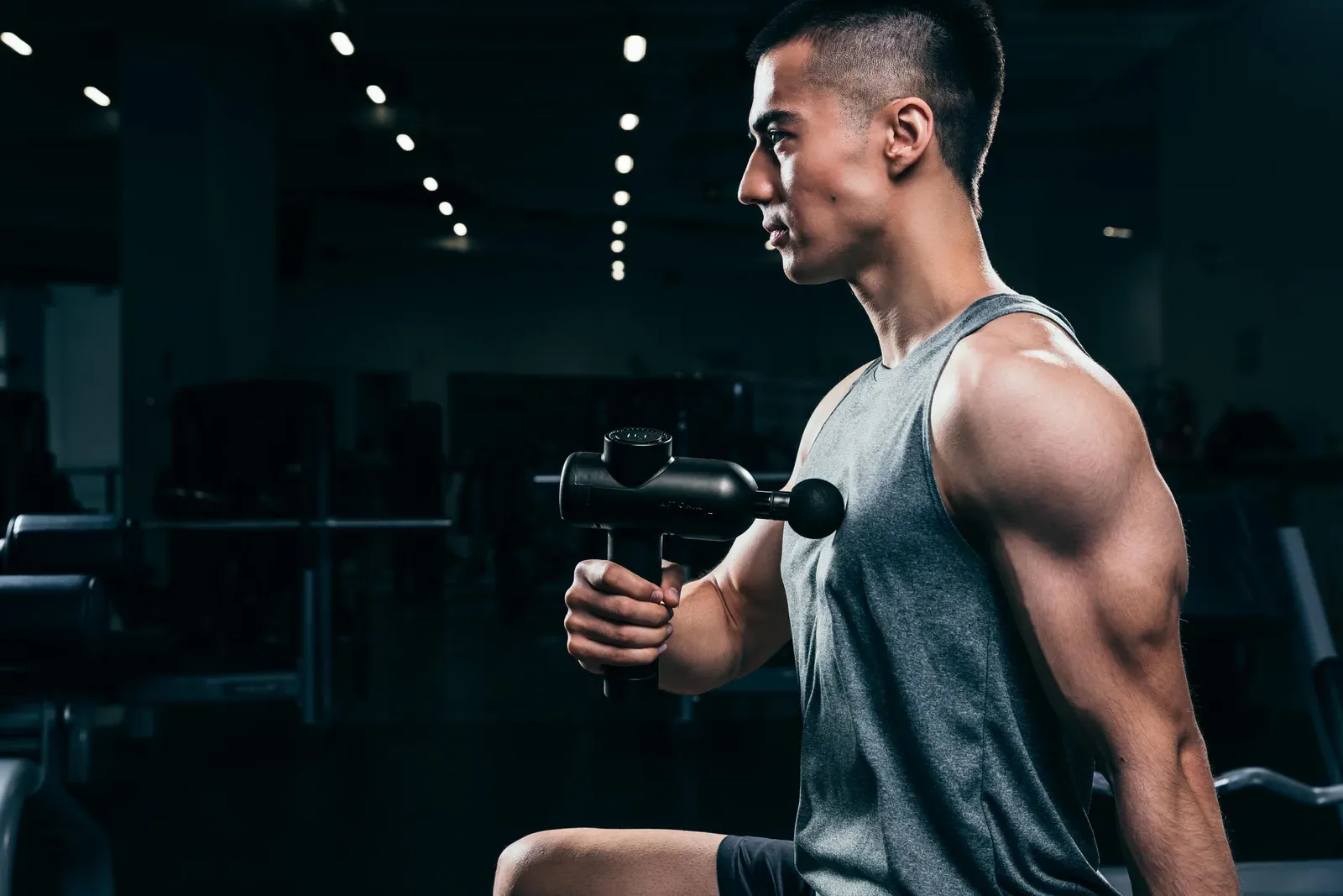 Man in a grey tank top using a ball head massager on his chest in a dimly lit gym