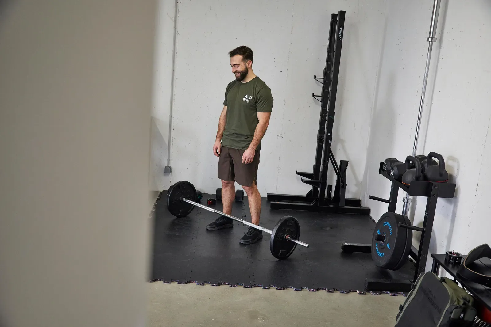Man standing over a loaded barbell on rubber floor tiles in a home gym with a rack