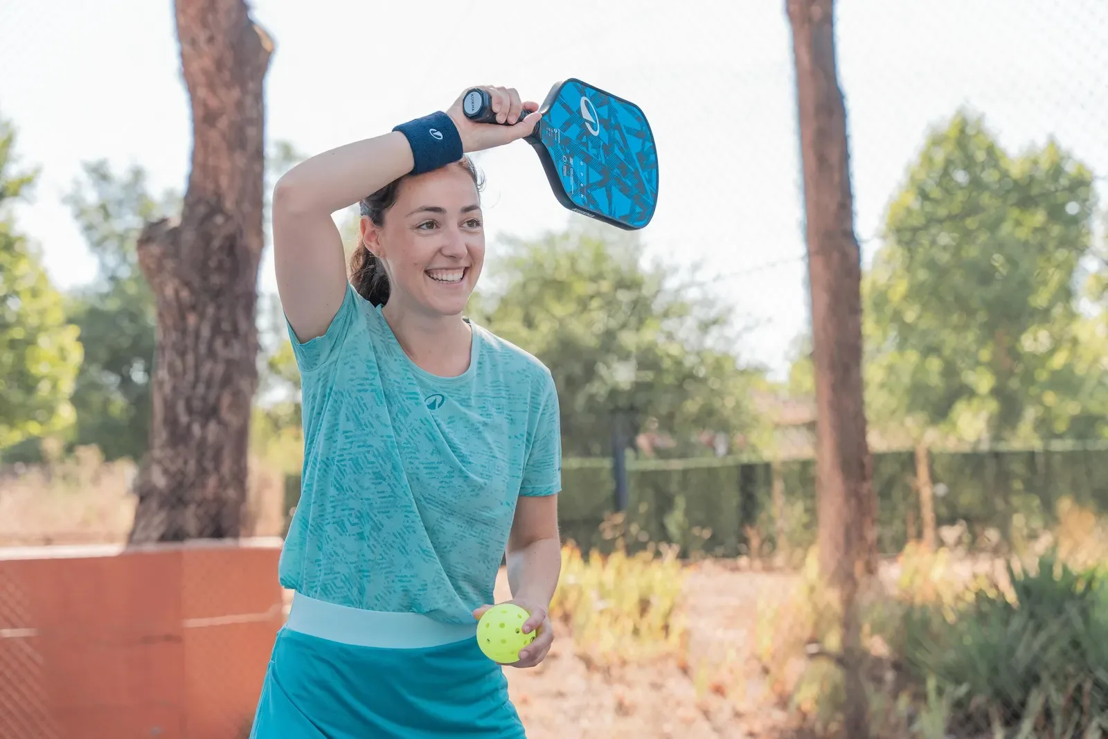 Smiling pickleball player holding a blue paddle and ball courtside