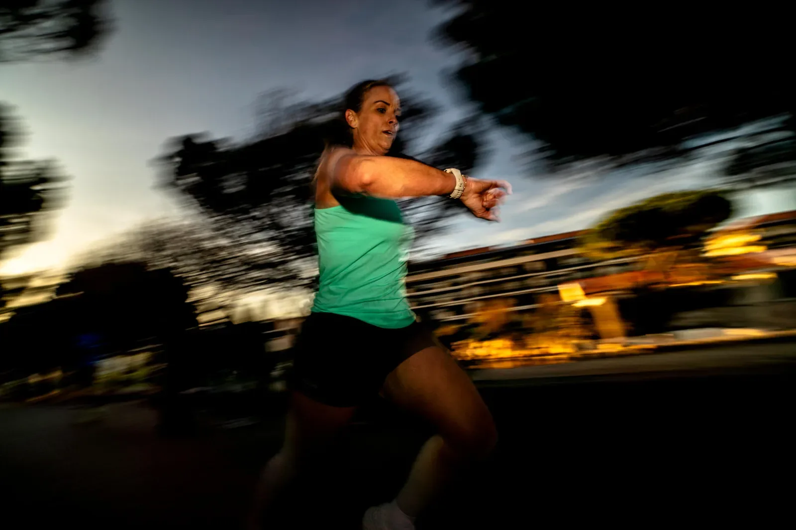 Woman checking her watch mid run in the evening with city lights blurred behind