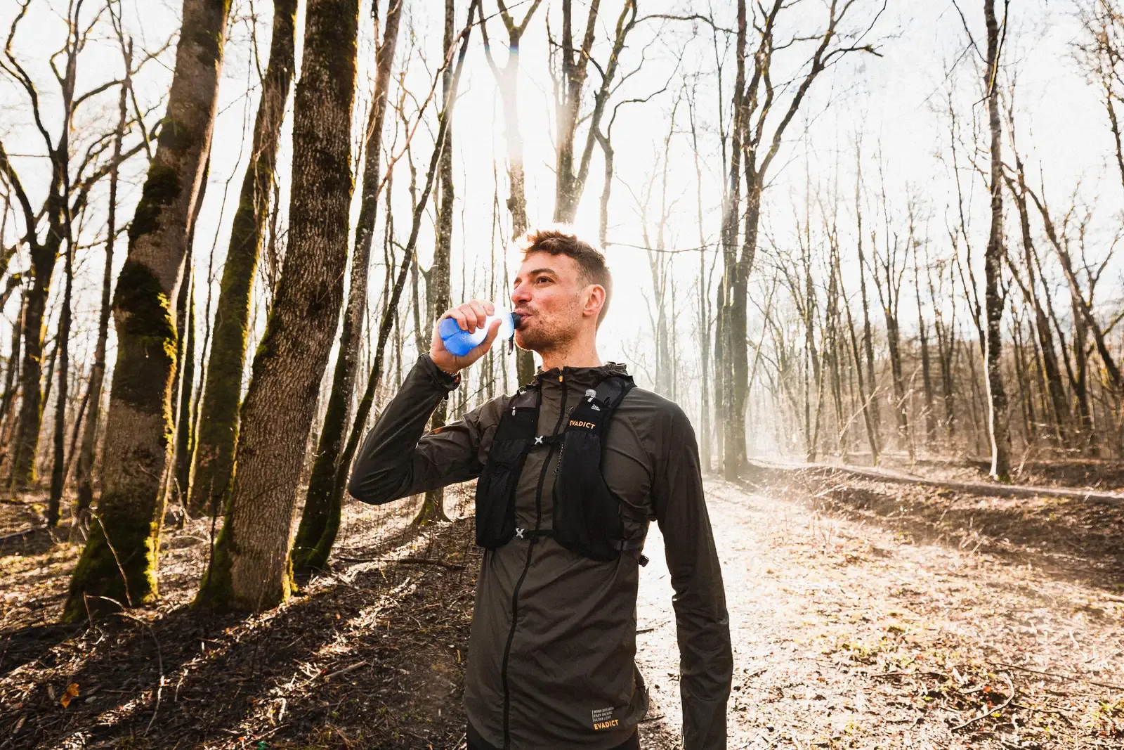 Runner in a hydration vest sipping from a soft flask in a sunlit forest