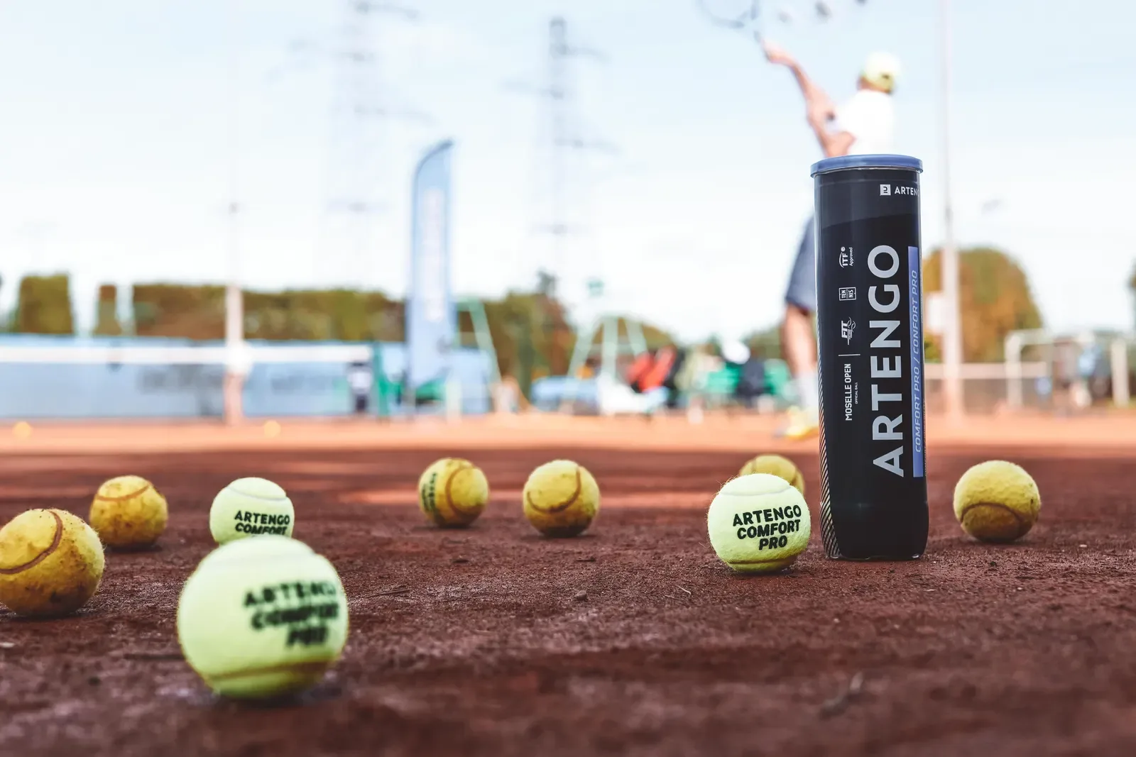 Tennis balls and a ball tube on a clay court with a player serving behind