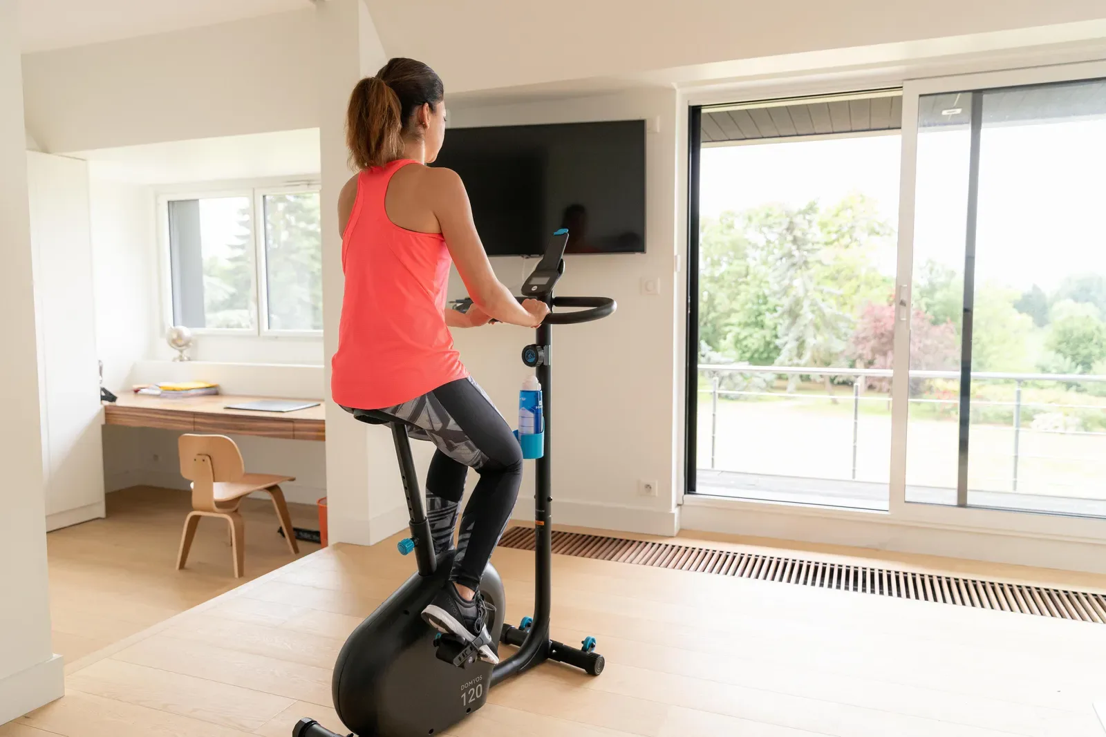 Woman riding an exercise bike at home with her desk and chair in the corner