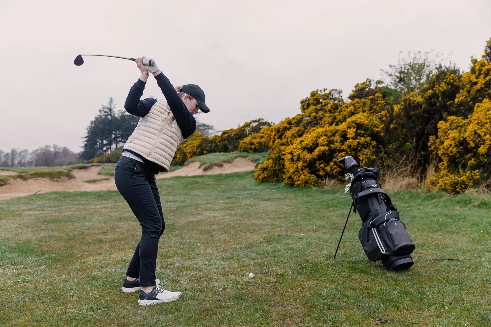 Woman in a padded vest at the top of her backswing on a coastal links course, golf bag on its stand