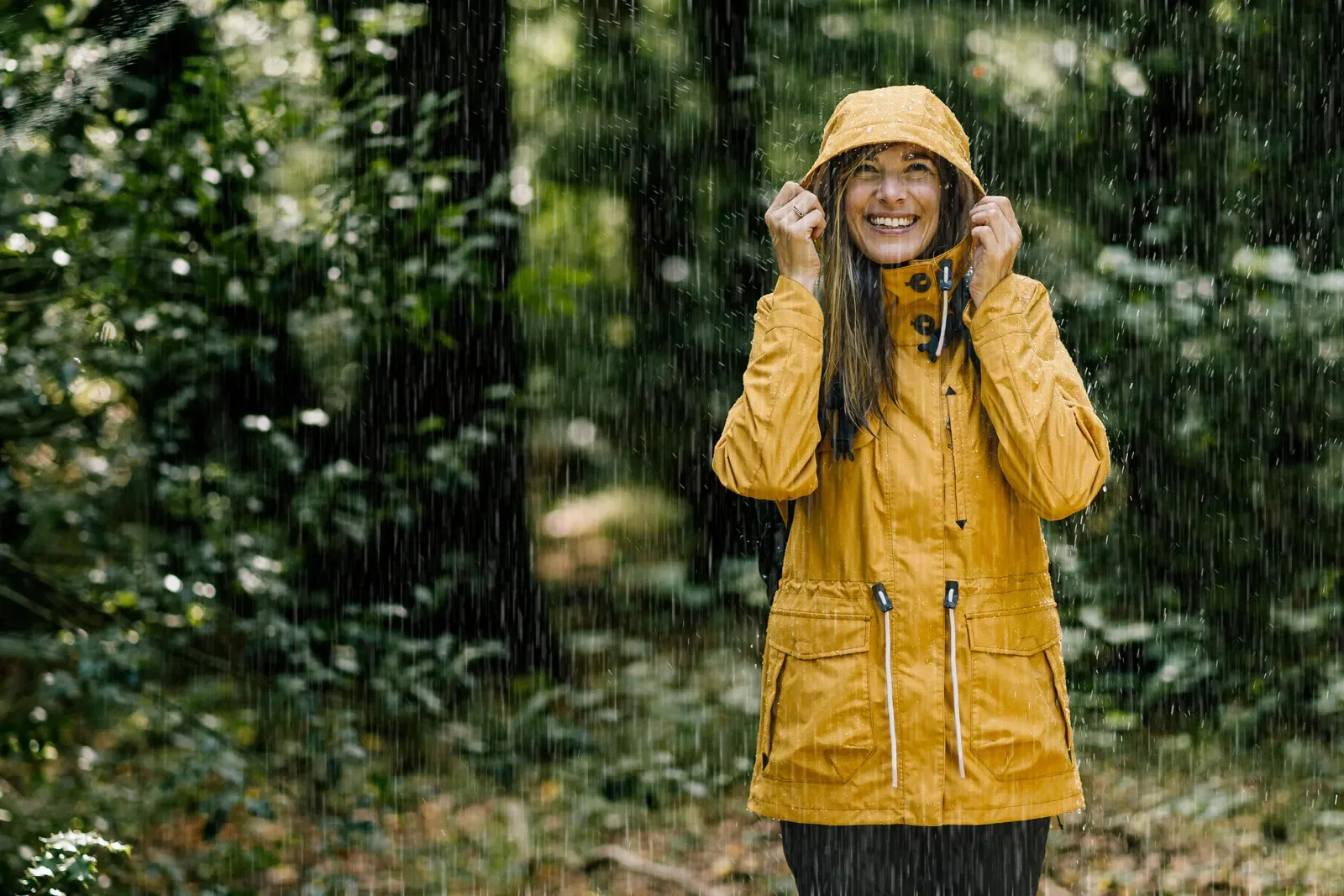 Smiling woman in a yellow rain jacket standing in the rain in a lush green forest